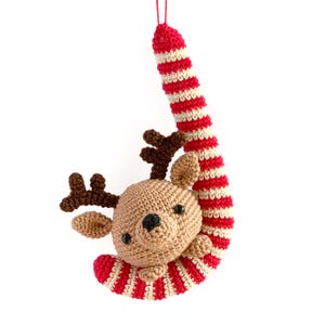 Puede incluir: Un adorno de reno de ganchillo colgado de un bastón de caramelo rojo y blanco. El reno es marrón con la nariz negra y las astas marrones.
