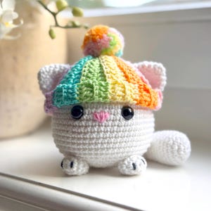 May include: A handmade white crochet cat with black safety eyes, a pink nose, and a rainbow-colored hat with a pom-pom. The cat has small ears and paws. The cat is sitting on a white surface.