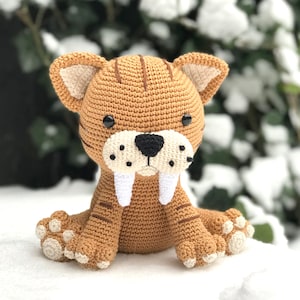 May include: A crocheted brown and white saber-toothed tiger plush toy with black eyes and a white mouth. The toy is sitting on a white surface.