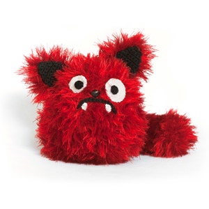 May include: A red fuzzy crocheted monster with black ears, white eyes, and a black nose. The monster has a wide, toothy grin and a long, fluffy tail.