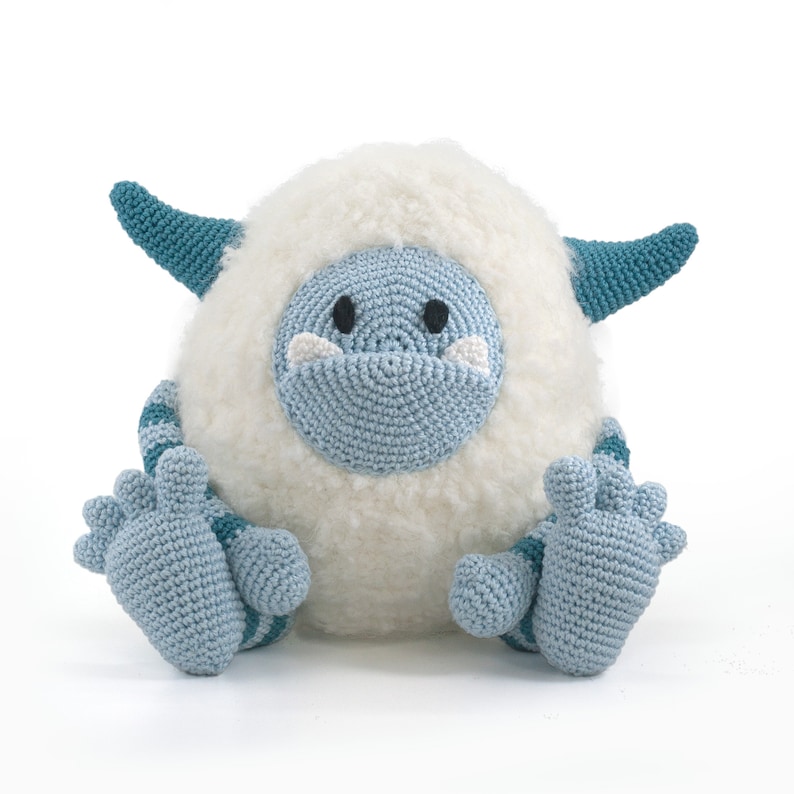 May include: A crocheted plush toy resembling a Yeti, with a white fluffy body, blue and teal arms and legs, and a blue face with black eyes.