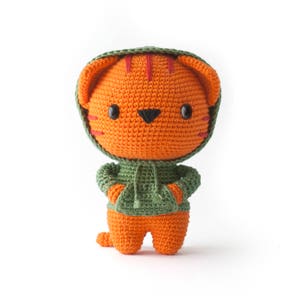 May include: A crocheted orange cat wearing a green hooded sweatshirt. The cat has black eyes and a black nose.
