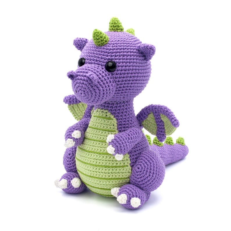 May include: A purple and green crocheted dragon toy. The dragon has a green horn, green wings, and white claws.
