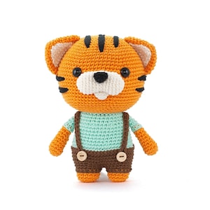 Puede incluir: Un juguete de peluche de tigre tejido a crochet que lleva una camisa azul claro y un mono marrón. El juguete tiene rayas negras y una cara blanca con ojos negros.
