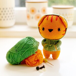 Pode incluir: Uma figura de tigre laranja em croch&ecirc; vestindo um moletom verde com capuz, posicionada ao lado de um novelo de l&atilde; verde, um novelo de l&atilde; laranja, uma pequena agulha de croch&ecirc; amarela e dois olhos de seguran&ccedil;a pretos com arruelas.