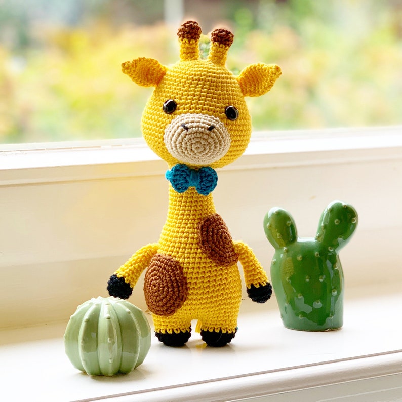 May include: A crocheted yellow giraffe toy with brown spots and a blue bow tie. The giraffe is standing on a white surface next to two green cactus-shaped decorations.
