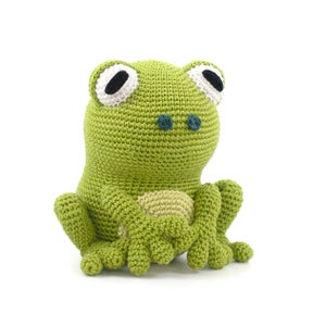 May include: A handmade green crochet frog with large white-rimmed eyes and black pupils. The frog has a light beige belly and is sitting upright with its arms and legs extended. The frog is made of soft yarn and is a cute, whimsical toy.