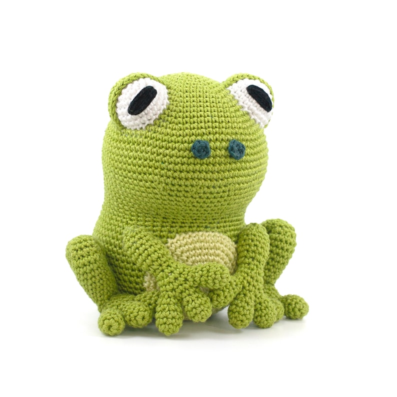 May include: A handmade green crochet frog with large white-rimmed eyes and black pupils. The frog has a light beige belly and is sitting upright with its arms and legs extended. The frog is made of soft yarn and is a cute, whimsical toy.