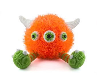 May include: A bright orange crocheted monster with three green eyes and white horns. The monster has green crocheted arms and legs.