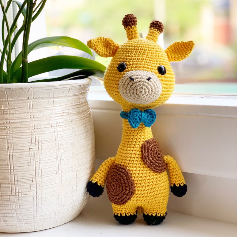 May include: A crocheted giraffe toy with brown spots, a blue bow tie, and black eyes. The giraffe is standing on a white surface.