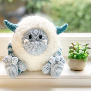 May include: A crocheted Yeti plush toy with blue and white fur, sitting on a windowsill. The Yeti has a blue face with black eyes and a white mouth.