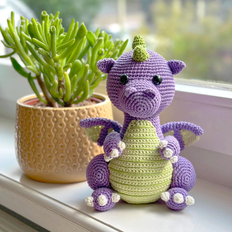 May include: A crocheted purple and green dragon toy with white claws. The dragon has a green horn and black eyes.