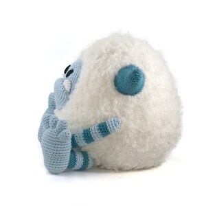 May include: A crocheted plush toy in the shape of a white yeti with blue and teal accents. The yeti has a large, fluffy body and a small, blue face with a single black eye. The yeti is sitting on its side with its arms crossed.