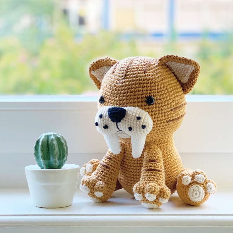 May include: A crocheted stuffed toy of a saber-toothed tiger, sitting on a windowsill. The toy is brown and white with black eyes and a black nose. It has large, white, pointed teeth.