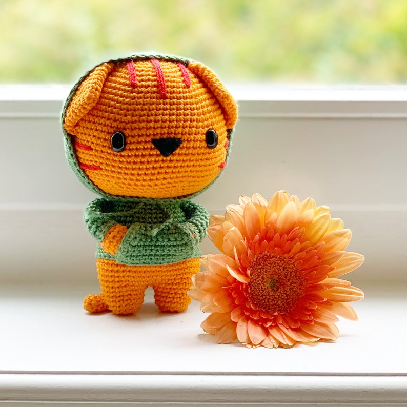 Pode incluir: Um peluche de tigre laranja feito em croch&eacute;, vestindo uma camisola verde com capuz e bolso frontal, com olhos de contas pretas e riscas vermelhas cosidas na cabe&ccedil;a, posicionado ao lado de uma grande flor gerbera laranja clara.
