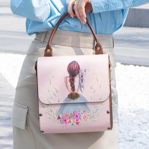 May include: Pink handbag with brown handles and trim. The front features a watercolour illustration of a girl with a koala and floral accents. The bag has a clasp closure and metal studs.