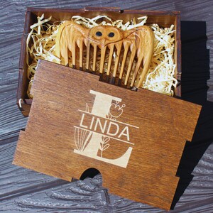 K&ouml;nnte beinhalten: Ein Holzkamm mit Eulen-Design, pr&auml;sentiert in einer Holzbox. Auf dem Deckel der Box ist der Name "LINDA" eingraviert. Der Kamm ist hellbraun, die Box ist dunkler braun. Die Box enth&auml;lt Holzwolle zur Pr&auml;sentation.