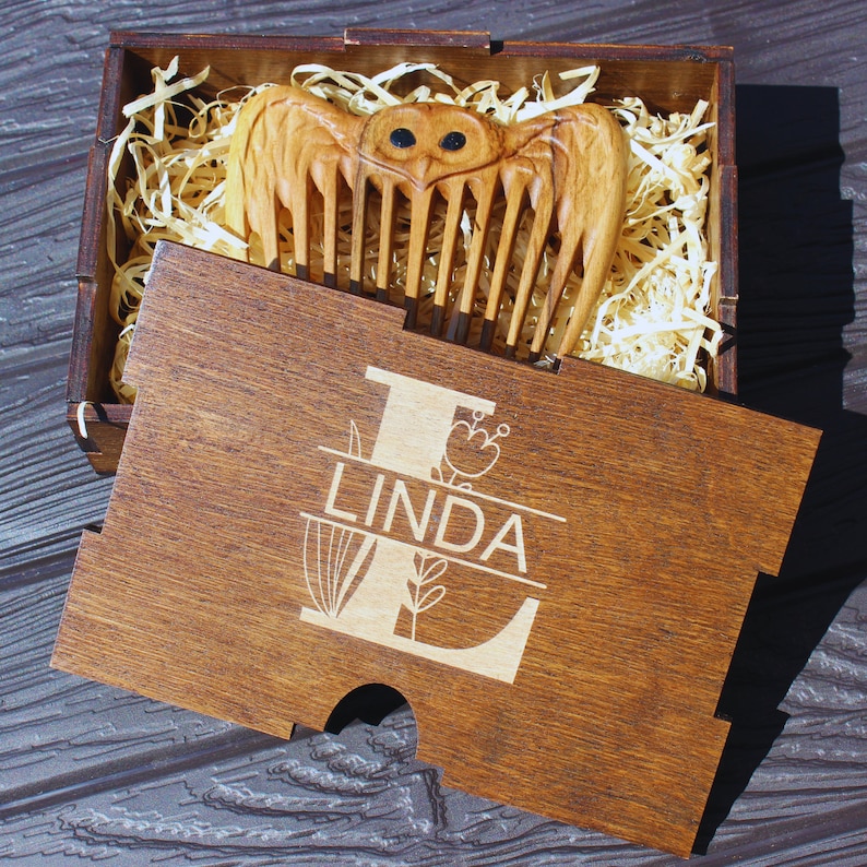 K&ouml;nnte beinhalten: Ein Holzkamm mit Eulen-Design, pr&auml;sentiert in einer Holzbox. Auf dem Deckel der Box ist der Name "LINDA" eingraviert. Der Kamm ist hellbraun, die Box ist dunkler braun. Die Box enth&auml;lt Holzwolle zur Pr&auml;sentation.