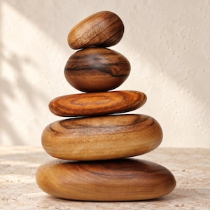 May include: A vertical stack of five smooth, polished wooden stones of varying sizes. Each piece features a distinct natural wood grain pattern with warm brown and tan tones, arranged from the smallest stone at the top to the largest at the base.
