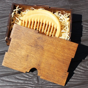 May include: A wooden comb in a wooden box with wood shavings. The comb is light brown and has a rounded shape. The box is dark brown and has a lid.