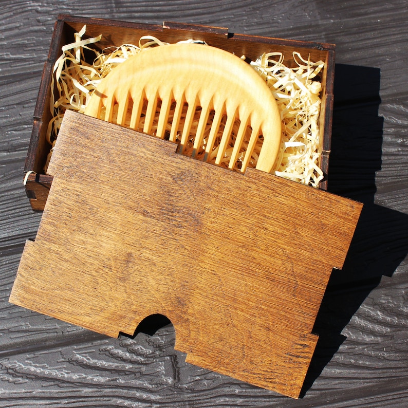 May include: A wooden comb in a wooden box with wood shavings. The comb is light brown and has a rounded shape. The box is dark brown and has a lid.