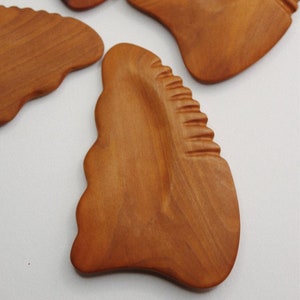 May include: A brown wooden gua sha tool with a textured surface. The tool is shaped like a leaf with a rounded edge.