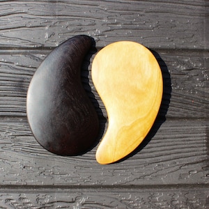 May include: Two wooden gua sha tools. One is dark brown, the other is light yellow. The tools are teardrop-shaped and rest on a dark, textured surface. The tools are designed for facial massage and skincare.
