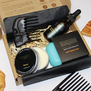 May include: A gift box set for beard care with a wooden comb shaped like a skull, a small tin of beard balm, a blue soap bar, a bottle of beard oil, and a second comb. The box is lined with shredded paper and the products are labeled with the brand name "BORODA".