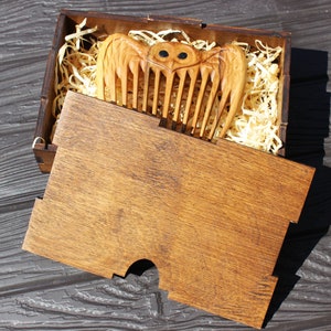K&ouml;nnte beinhalten: Ein Holzkamm mit Eulenmotiv, pr&auml;sentiert in einer Holzbox mit Schiebedeckel. Der Kamm ist hellbraun mit geschnitzten Details, einschlie&szlig;lich der Fl&uuml;gel und Augen der Eule. Die Box ist mit Holzsp&auml;nen gef&uuml;llt.
