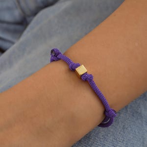 Pode incluir: Um bracelete de cordão roxo com um fecho quadrado dourado.