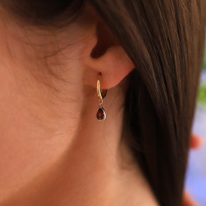 Könnte beinhalten: Goldene Creolen mit einem roten Edelstein, der herunterhängt. Die Ohrringe werden am Ohr einer Person getragen.