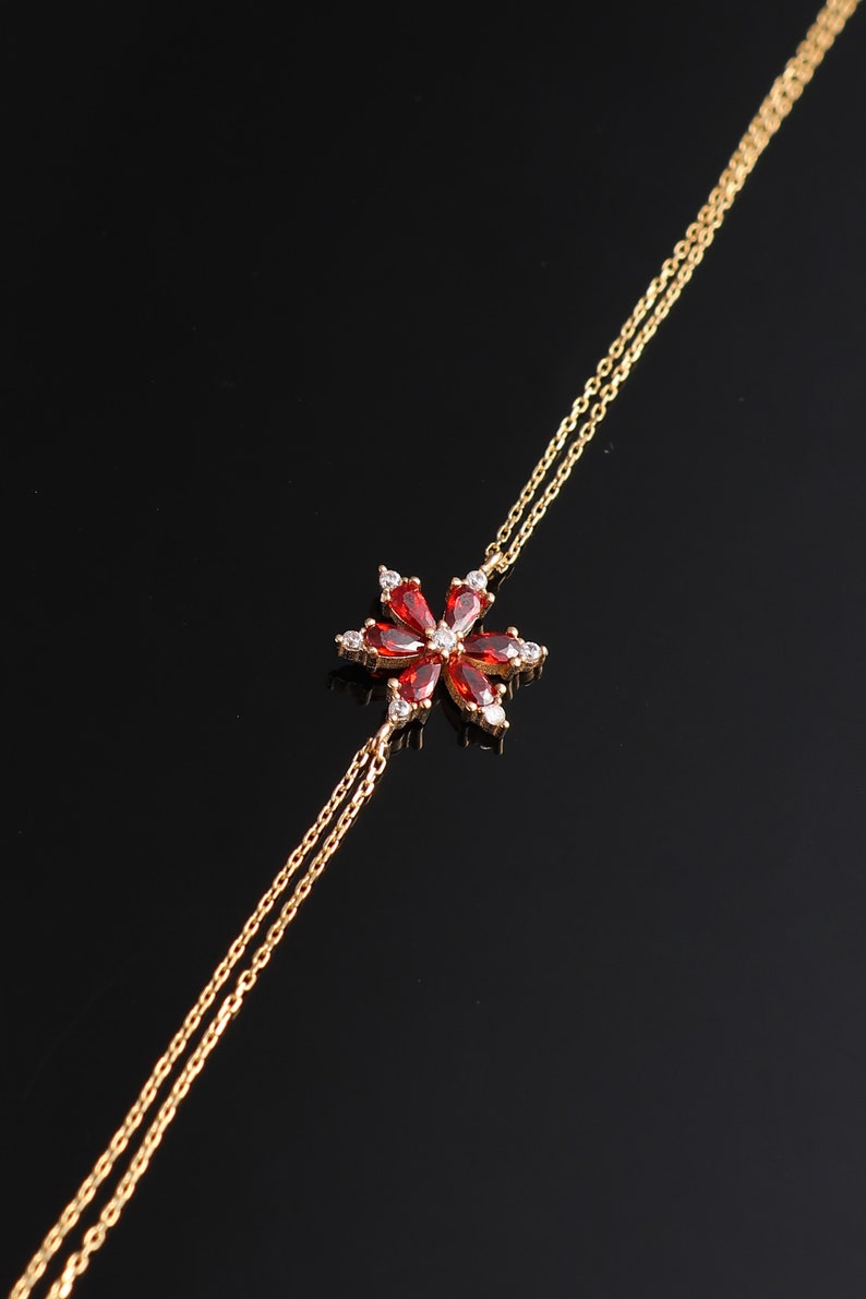 May include: A gold chain bracelet with a red and white flower charm. The flower has a five-petal design with clear stones in the center of each petal.