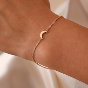 May include: A delicate gold-colored bracelet featuring a crescent moon charm. The bracelet is made of a fine chain and is worn on a wrist. The moon charm is centered on the chain, adding a touch of celestial elegance to the jewelry.