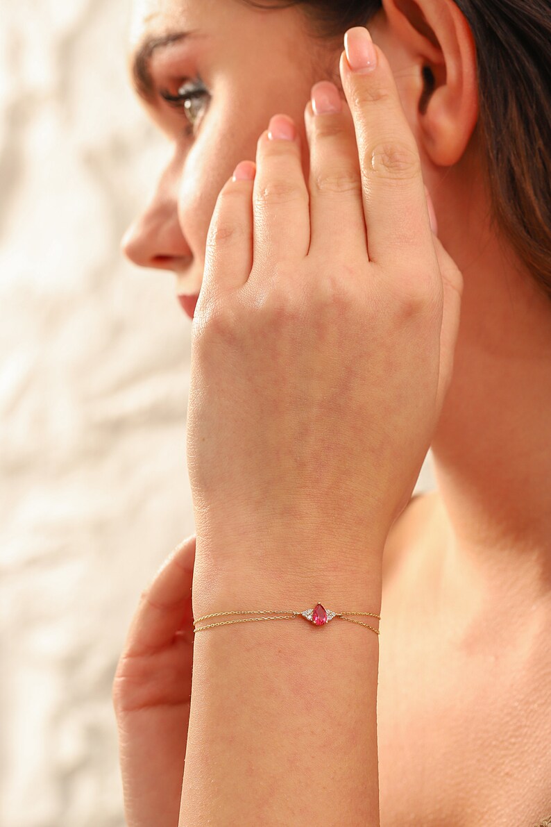 May include: A delicate gold chain bracelet with a pear-shaped pink gemstone and two smaller white gemstones.