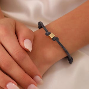 Pode incluir: Uma pulseira trançada cinza com um fecho quadrado dourado.