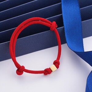 Pode incluir: Um bracelete de corda vermelha com um fecho quadrado dourado. O bracelete é amarrado com um nó em cada extremidade.