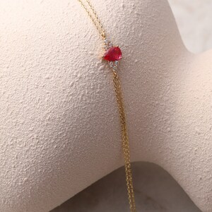 May include: A delicate gold chain necklace with a teardrop-shaped red gemstone pendant surrounded by smaller clear gemstones.