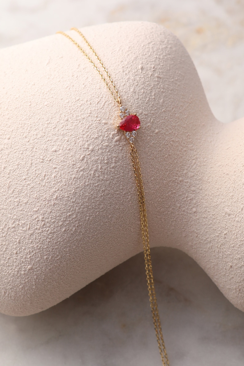 May include: A delicate gold chain necklace with a teardrop-shaped red gemstone pendant surrounded by smaller clear gemstones.