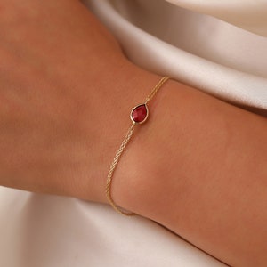 May include: A gold chain bracelet with a teardrop-shaped red gemstone pendant.
