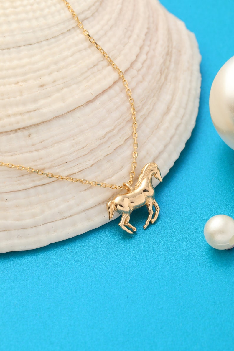 May include: A gold chain necklace with a horse pendant. The necklace is on a white seashell and a blue background.