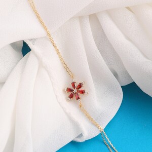 May include: A delicate gold chain necklace with a red gemstone flower pendant. The flower has a center stone surrounded by smaller stones.