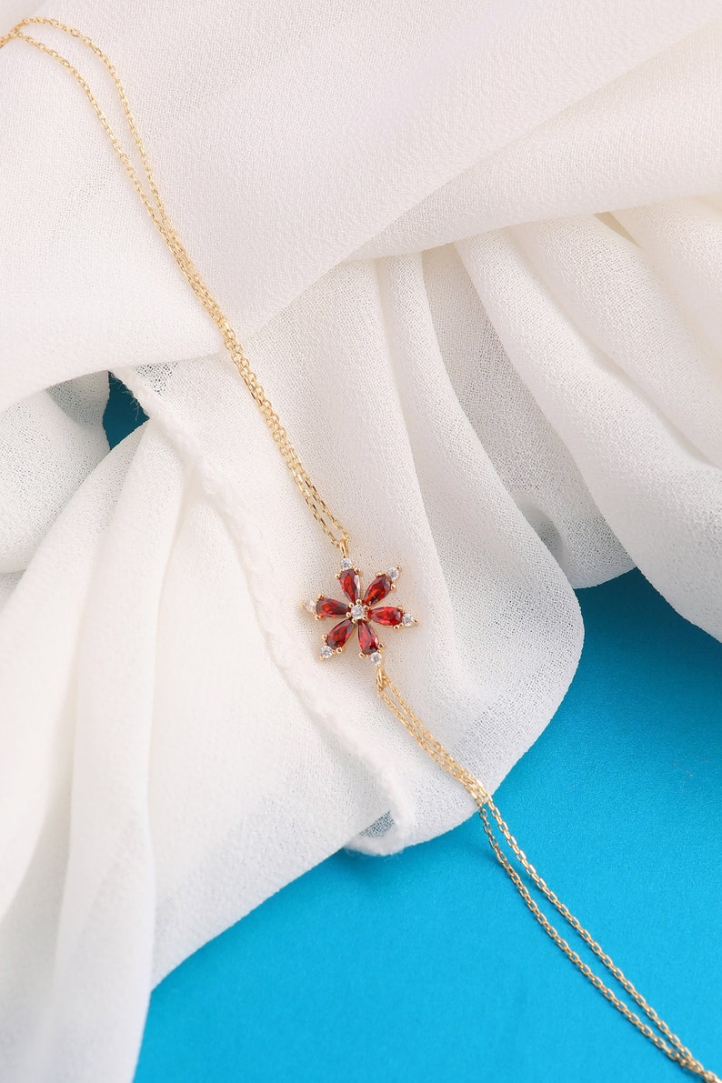 May include: A delicate gold chain necklace with a red gemstone flower pendant. The flower has a center stone surrounded by smaller stones.
