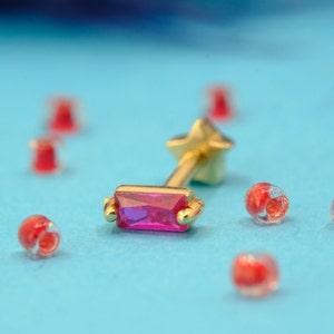 May include: A gold-colored stud earring with a rectangular, magenta-colored gemstone. The earring has a star-shaped backing. Small, red and clear beads are scattered on a light blue surface.