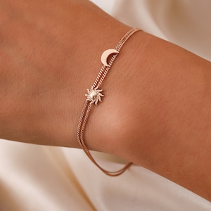 Puede incluir: Un delicado brazalete de cadena dorada con un dije de sol y luna. El brazalete está hecho de dos cadenas finas y presenta un pequeño dije de sol en una cadena y un dije de luna creciente en la otra.