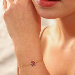 May include: A delicate gold chain bracelet with a red gemstone flower charm. The flower charm is made of small red gemstones set in a gold setting. The bracelet is a simple and elegant piece of jewelry.