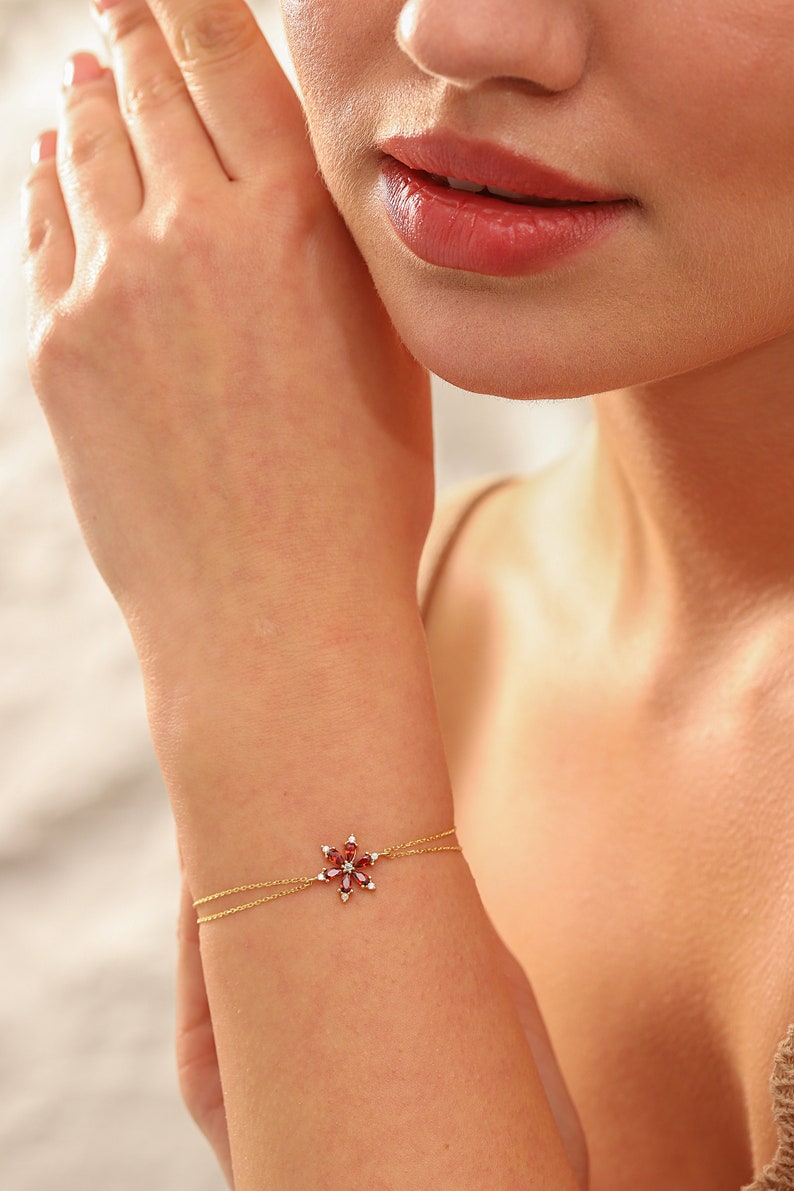 May include: A delicate gold chain bracelet with a red gemstone flower charm. The flower charm is made of small red gemstones set in a gold setting. The bracelet is a simple and elegant piece of jewelry.