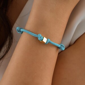 Pode incluir: Um bracelete de corda azul turquesa com um fecho quadrado dourado.