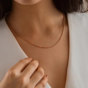 May include: A delicate gold chain necklace with a beaded design. The necklace is worn by a person with a white shirt.
