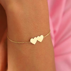 Op de afbeelding: Een delicate gouden armband met twee hartvormige bedels. De ketting van de armband heeft een fijn, getextureerd ontwerp. De armband wordt gepresenteerd tegen een roze achtergrond, wat de warme, gouden tint van de sieraden benadrukt.