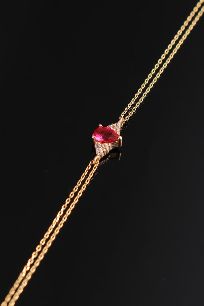 May include: A delicate gold chain necklace with a red gemstone pendant surrounded by smaller clear gemstones.